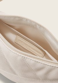 Intérieur d'un sac en toile beige avec doublure lisse, fermeture à glissière et une petite étiquette en tissu indiquant "DENIM TOM TAILOR". Surface extérieure texturée.