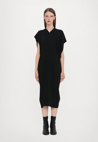 Robe noire longueur genoux avec un décolleté en V et des manches courtes. Tissu lisse avec une coupe décontractée, accentuée par des bottes noires hauteur cheville.