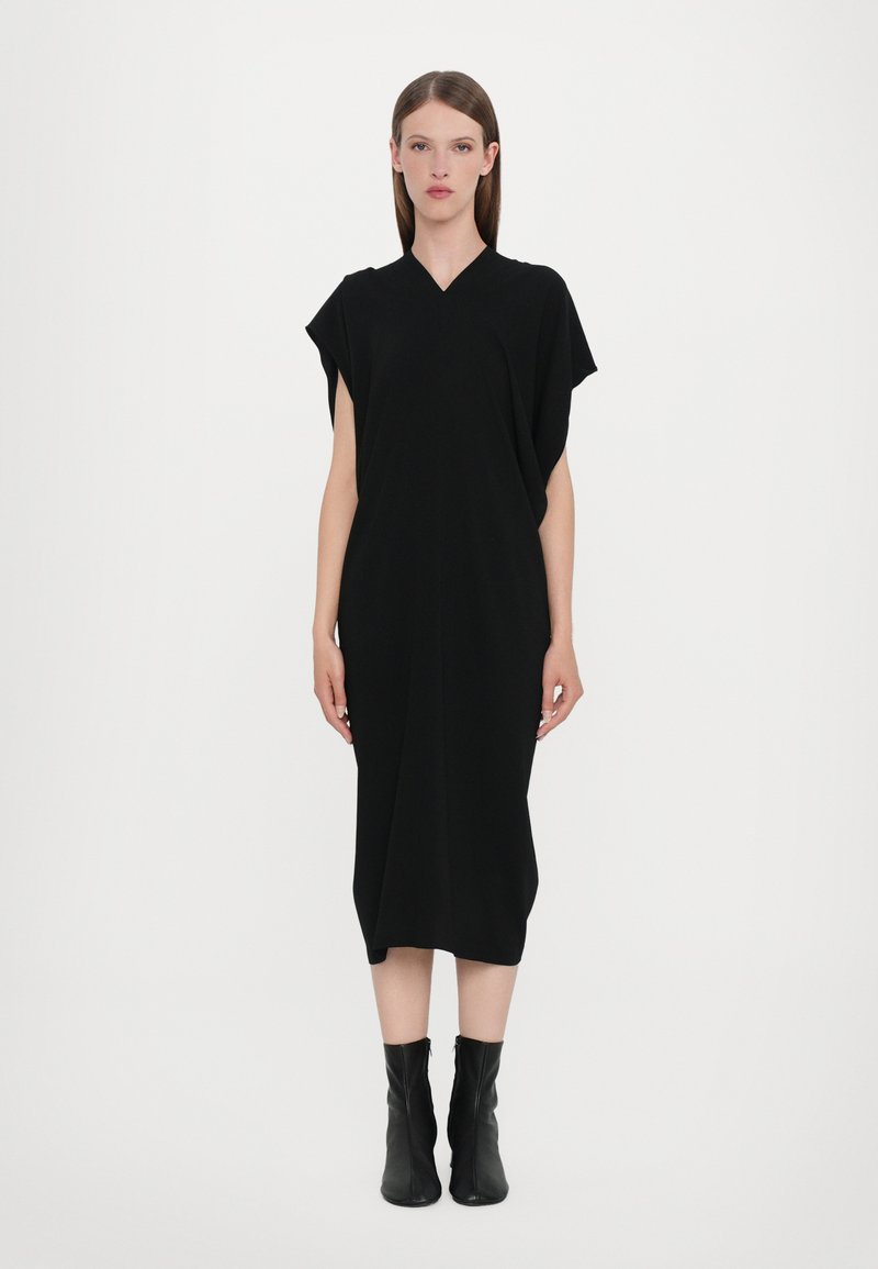 Black knee-length dress with a V-neck and short sleeves. Smooth fabric with a relaxed fit, accented by ankle-height black boots.