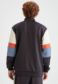 Veste noire avec des manches en coloris crème, orange et bleu ; dotée d'un col montant et de poignets côtelés, confectionnée dans un tissu lisse.