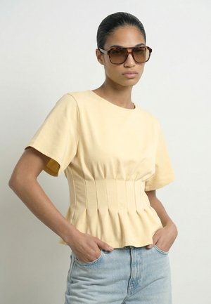Femme portant un top beige à manches courtes avec des plis et un jean bleu clair, posant les mains dans les poches et portant de grandes lunettes de soleil marron.
