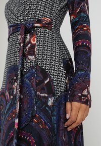 Nahaufnahme einer Hand, die an der Seite ruht, bekleidet mit einem gemusterten Langarmkleid mit geometrischen und kreisförmigen, bunten Mustern, das in der Taille gebunden ist.