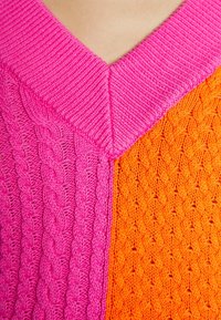 Dzianinowy sweter z dekoltem w kształcie litery V, podzielony na dwie części: jasnoróżową z warkoczowym splotem i jaskrawo pomarańczową z teksturą w diamentowy wzór.