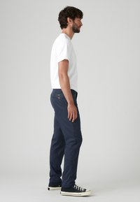 Pantaloni marini cu croială dreaptă, dintr-un material ușor, cu buzunare laterale și o patch logo subtil pe spate. Modelul poartă un tricou alb și încălțăminte neagră.