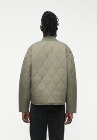 Asmuo su tamsiais pintais plaukais, vilkintis alyvuogių žalią dygsniuotą striukę ir juodas kelnes, stovintis nugara priešais paprastą baltą foną.