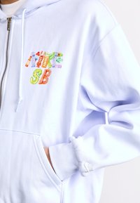 Lichtblauwe hoodie met rits en meerkleurige geborduurde tekst. Het materiaal is zacht, met een zak aan de voorkant en geribbelde boorden.