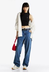 Sort crop top, hvid skjorte med knapper, oversized blå jeans, sort bælte med logo, rød håndtaske og cremefarvede sneakers med brune detaljer.