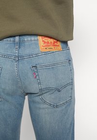 Modré džínové kalhoty s vysokým pasem, s kapsami vzadu, viditelnou červenou etiketu a oranžovou koženou značkou s motivem kovboje.