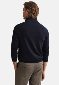 Mörkblå stickad tröja med hög krage och ribbade muddar, kombinerad med texturerade bruna byxor med två knappar bak.