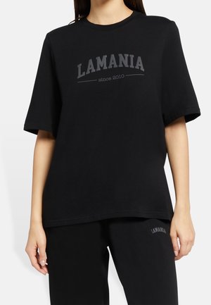 Femme portant un T-shirt noir ample et un pantalon noir, tous deux avec le mot "LAMANIA" imprimé sur le devant, debout devant un fond uni.