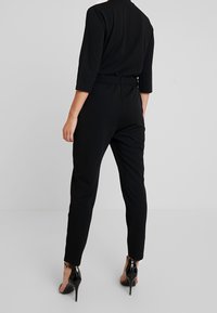 Mono negro con mangas tres cuartos, diseño ajustado, detalle en la cintura y piernas entalladas, combinado con zapatos de tacón alto negro brillante.
