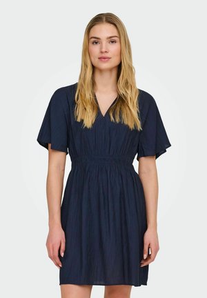 Ble blondinka z razpuščenimi lasmi, oblečena v kratko, temno modro obleko z nabranim pasom, stoji pred enobarvnim svetlim ozadjem.