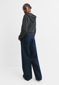 Felpa con cappuccio grigio scuro dal taglio relaxed, abbinata a jeans in denim blu scuro a gamba ampia con cuciture visibili e dettagli delle tasche.