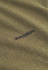 Olivno zelena tkanina z gladko teksturo, na kateri je temno moder napis »GABBA«. Dizajn je preprost in moden ter poudarja funkcionalnost.