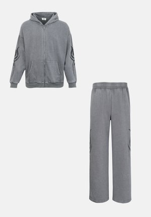 Šedá mikina s kapucí na zip a široké kalhoty v odpovídajícím stylu s černými kmenovými motivy na rukávech a nohavicích kalhot.