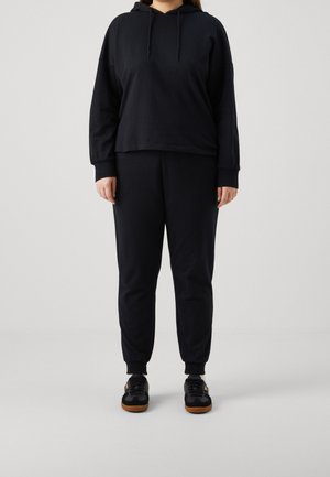 Person trägt schwarzen Hoodie mit langen Ärmeln und passende schwarze Jogginghose mit schwarzen Sneakers mit braunen Sohlen, steht vor einem einfarbigen hellen Hintergrund.