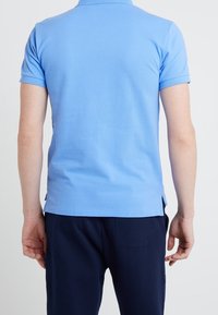 Ljusblå polo med mjuk textur, korta ärmar och sidokilar. Bakifrån visar den en ren design utan synliga logotyper eller detaljer.