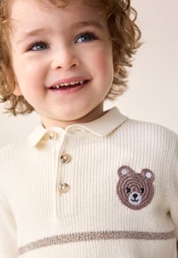 Kremowa dzianinowa koszulka polo o prążkowanej fakturze, z brązowymi poziomymi paskami i haftem twarzy niedźwiedzia na piersi. Trzy guziki.