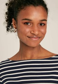 Čierno-biele pruhované tričko s širokým výstrihom, nosené osobou s kučeravými vlasmi. Obsahuje malé kruhové náušnice a hladkú pleť.