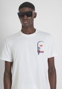 Camiseta de algodón blanca con un cuello redondo que presenta un diseño gráfico en el lado izquierdo del pecho, que incluye hojas de palma y rayas de colores.