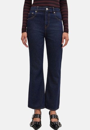 Jeans blu scuro a vita alta e gamba dritta indossati con scarpe nere a punta con grandi fibbie e dettagli con occhielli.