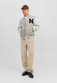Grijze en witte varsityjack met een "N"-insigne, drukknoopsluitingen, ribgebreide manchetten; gecombineerd met beige broeken en zwarte schoenen. Bruine pet gedragen.