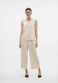 Beige, ärmlös blus med krage och framknappar, tillsammans med vida beige byxor, som visar ett mjukt, texturerat tyg.