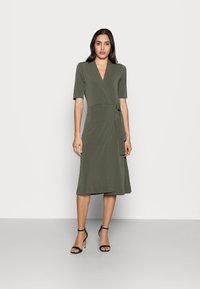 Vestido envolvente verde oliva com mangas curtas, decote em V e laço na cintura. Tem uma bainha até ao joelho e uma textura suave. Sandálias de salto alto pretas.