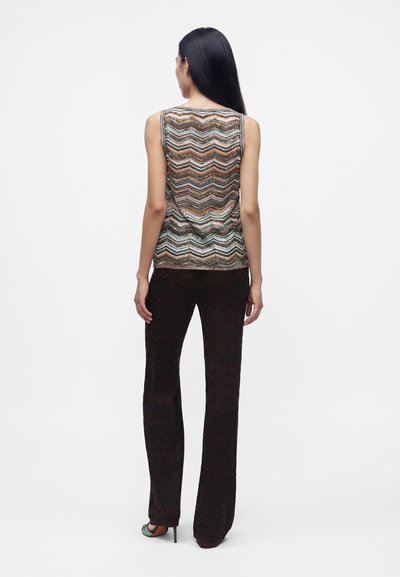 Femme aux longs cheveux noirs portant un haut sans manches à motif chevron et un pantalon noir à jambes larges, debout de dos sur un fond blanc.