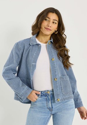 Femme aux longs cheveux ondulés portant des boucles d'oreilles créoles dorées, un haut blanc, une veste à boutons rayée bleu et blanc, et un jean bleu clair, posant avec la main dans la poche.