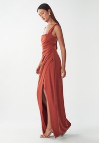 Langes, rostfarbenes Satin-Kleid mit quadratischem Ausschnitt, seitlicher Raffung und hohem Schlitz; getragen mit nude-farbenen Riemchensandalen und goldenen Ohrringen.