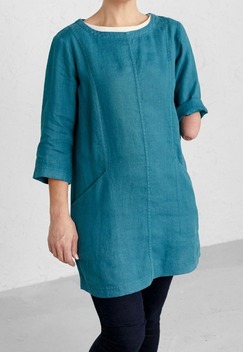 Personne avec un avant-bras gauche manquant, portant une tunique bleu sarcelle à manches longues et un pantalon noir, se tenant devant un fond clair uni.