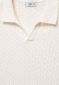 Krémově zbarvené pletené polo tričko se strukturovaným vzorem, s plochým límcem a výstřihem do V. Na krku je viditelná etiketa.