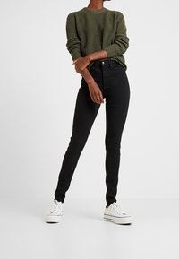 Svarta smala jeans i kombination med en texturerad grön stickad tröja. Outfiten fulländas av vita sneakers och har en figurnära silhuett.