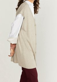 Beige stickad väst med knappar, med avslappnad passform och djupa ärmhål, lagd över en vit skjorta med uppvikta ärmar.