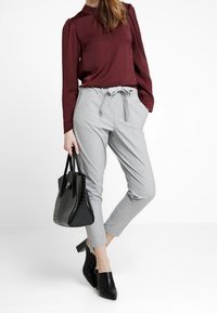 Pantalons gris ajustés avec une taille à nœud papillon, associés à un haut bordeaux à manches longues. Des chaussures à talons noires et un sac à main noir texturé complètent la tenue.