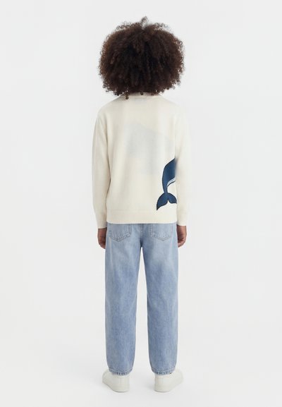 Personne aux cheveux bouclés vue de dos portant un pull blanc avec un motif de queue de baleine bleue et un jean bleu clair.