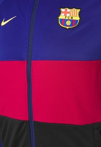 Nike zip-up jacket featuring blue, red, and black panels. Includes a Barcelona logo on the chest and smooth fabric texture.