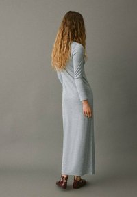 Abito maxi grigio a maniche lunghe con una silhouette aderente, morbido al tatto e con scollo rotondo. Abbinato a sandali marroni.