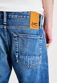 Bakifrån syns en person som bär blå jeans med en beige läderlapp märkt "DENHAM" som visar en saxlogotyp ovanför bakfickan.