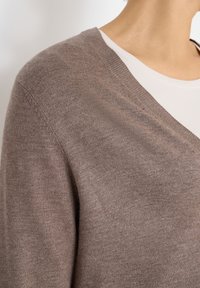 Bruine v-hals trui in een zachte, gestructureerde breisel met een ontspannen pasvorm. Gelaagd over een lichtgekleurde t-shirt, wat de halslijn accentueert.