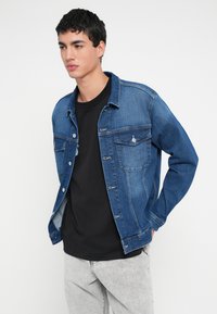 Junger Mann trägt eine blaue Jeansjacke über einem schwarzen Hemd und hellgrauen Jeans, steht mit einer Hand in der Tasche vor einem einfarbigen Hintergrund.