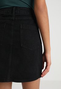 Černá džínová sukně se pevně střiženým pasem, dvěma zadními kapsami a rovného lemu. Dokončena tónovým prošíváním a jemnou texturou.