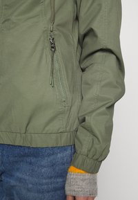 Olivengrøn bomberjakke med lynlåslomme, elastiske manchetter og ribbet kant. Har synlige syninger og et glat, tekstureret stof.