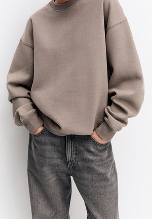 Person iført en løs taupe sweatshirt med ribbestrikkede manchetter og falmede sorte jeans, stående med hænderne i lommerne mod en ensfarvet baggrund.