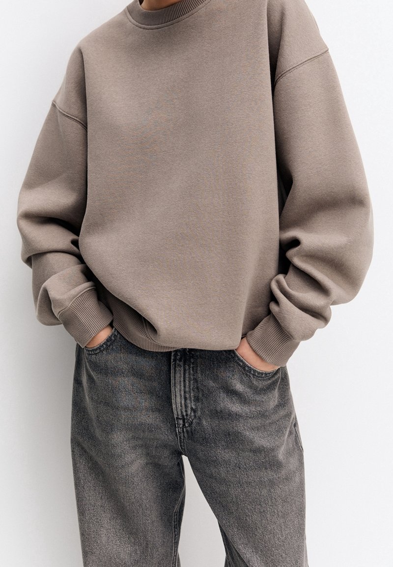 Personne portant un sweat-shirt ample taupe avec des poignets côtelés et un jean noir délavé, debout les mains dans les poches devant un fond uni.