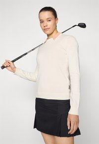 Κρέμ μακρυμάνικο πουλόβερ με ανοιχτό ροζ γιακά, ριχτή γραμμή και ριμπ ύφανση στο τζάκι, συνδυασμένο με μαύρη πλισέ φούστα, κρατώντας ένα μπαστούνι του γκολφ.