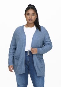 Világoskék kardigán hosszú ujjal, nyitott elülső résszel, két elülső zsebbel és bordázott mandzsettával. Fehér felső és kék farmert viselnek alatta.