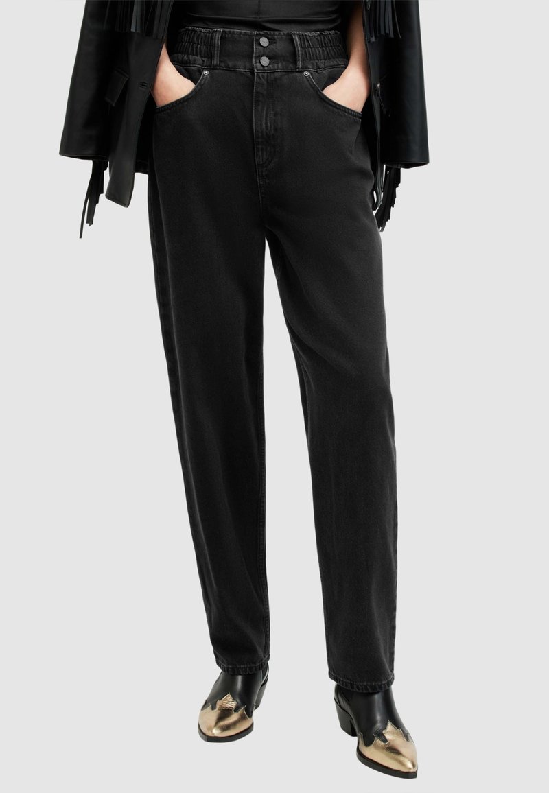 Pantalon en denim noir avec taille élastique, doté de poches latérales, d'une coupe ample, accompagné de bottes noires ornées d'accents dorés.