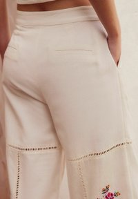 Pantaloni beige a gamba larga con vita alta, caratterizzati da ricami sottili, cuciture decorative lungo le cuciture e tasche posteriori. Texture del tessuto morbida.